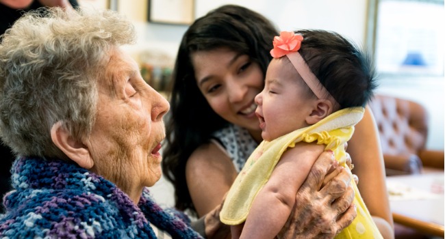 elderly-woman-holding-infant-granddaughter-as-mother-looks-on-smiling-picture-id541592296